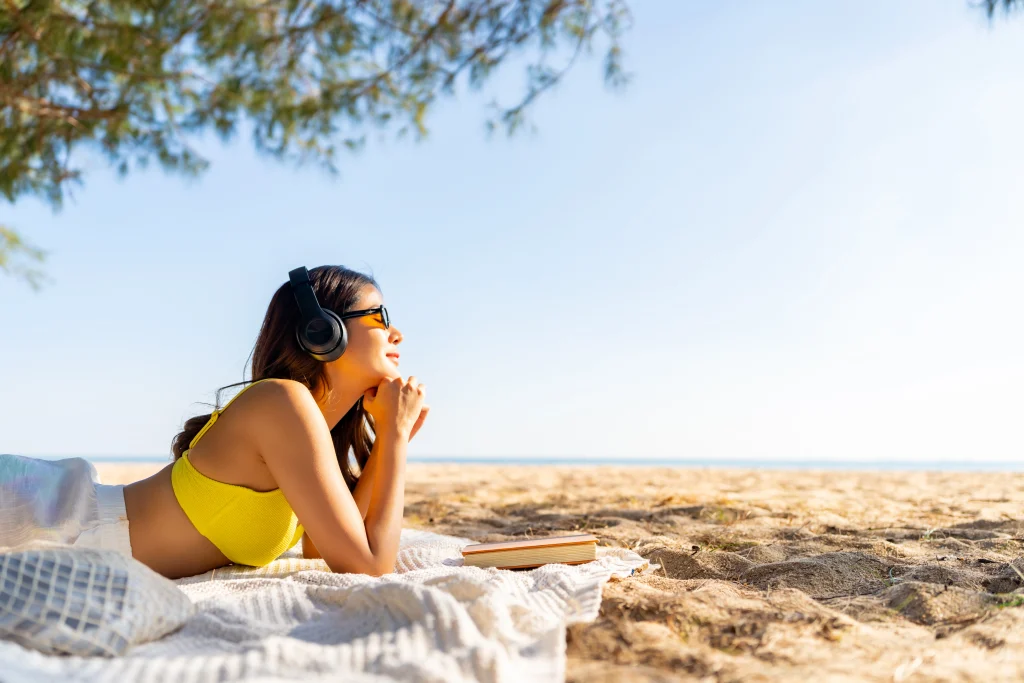 Femme allongée sur la plage avec des écouteurs, profitant d’une application de relaxation pour un moment de détente estivale.