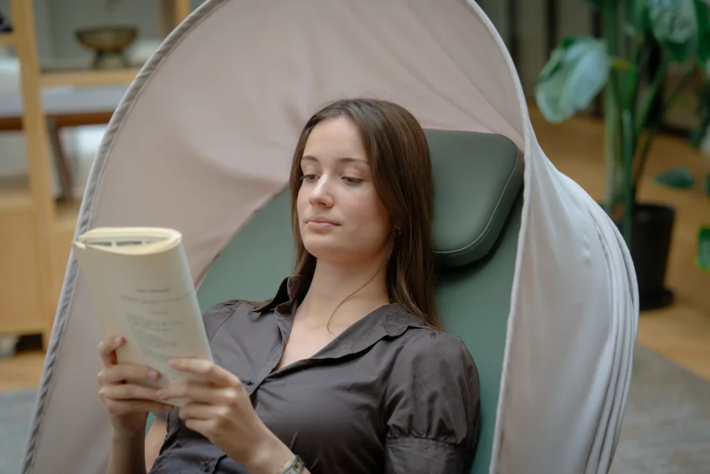 Une jeune femme brune lit un livre en position semi-allongée dans un fauteuil de relaxation. Le siège possède un appui-tête vert et une grande capote beige qui l'enveloppe. En arrière-plan, on aperçoit un intérieur chaleureux avec un parquet en bois et une plante d'intérieur.